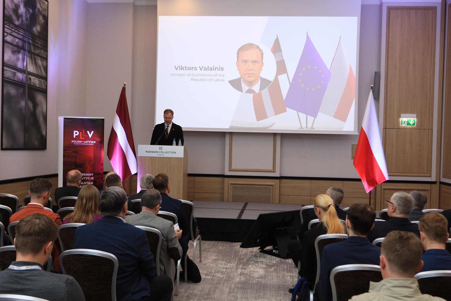 Ekonomikas ministrs Viktors Valainis Latvijas–Polijas biznesa forumā akcentē stratēģisko sadarbību drošības, tehnoloģiju un enerģētikas jomās Uz skatuves ekonomikas ministrs Viktors Valainis. Balkus skatuvei labajā malā Polijas karogs, kreisajā malā Latvijas karogs.