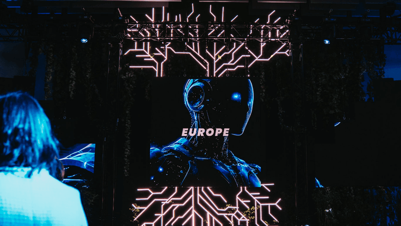Foto no Deep Tech Atelier 2026, kur uz melna ekrāna redzams zils robota siluets, kam fonā redzamas stilizētas mikroshēmas dzīslas no lejas u